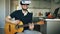 Happy young man sitting at kitchen learning to play guitar using VR 360 headset and feels him guitarist at concert at