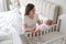 Happy young mother with her cute newborn baby near bedside crib at home