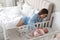 Happy young mother near bedside crib with cute newborn baby at home