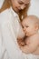 Happy young mother in white shirt breastfeed infant baby on white background closeup. Portrait of pretty naked little