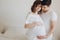 Happy young pregnant couple holding belly bump on white bed. Stylish pregnant family, mom and dad in white, relaxing at home and