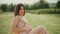 Happy young pregnant woman in a pink dress sitting among green plants and wildflowers. She gently strokes her stomach