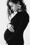 Happy young pregnant woman in stylish black dress and modern earring, holding belly bump and posing in light. Fashionable mom,