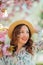 Happy young woman 30 years old in a wicker hat walks in a blooming city park. Portrait of a stylish emotional model in a light