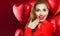 Happy young woman with heart balloons on red background. Surprised girl with red lips makeup and opened mouth.