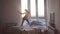 Happy young woman jumps and dances on bed in luxury apartment during beautiful sunrise with lens flare effect in the