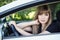 Happy young woman posing in her car holding steering wheel. Lady driver with long blond healthy hair at vehicle. Warm season