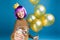 Happy young woman with purple haircut, crown on head celebrating with golden balloons and champagne on blue background