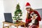 Happy young woman in santa hat sitting on bed with cat, dancing and looking at laptop. Online Christmas and new year celebration