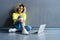 Happy young woman sitting on the floor with crossed legs and using laptop on gray background.