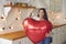 Happy young woman standing in the kitchen, holding huge Valentine balloon and smiling