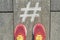 Hashtag symbol on gray sidewalk with woman legs in sneakers, top view