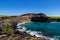 Hawaii`s green sand beach. Steep cliff, blue-green water, blue sky. Rocky shoreline in foreground.