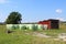 Hay bales wrapped in plastic next to wooden garage surrounded with uncut grass and large trees with family houses in background