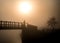 Hazy orange misty sunrise with lone man silhouette standing on river bridge looking at view of sun in fog. Stood wooden footbridge