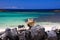 Head of blonde woman with straw hat sitting behind wall of piled natural rocks on beach with turquoise ocean - Fuerteventura, El