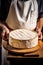 A head of brie cheese in man\\\'s cheesemaker hands. delicious dairy products. cheese camembert on wooden plank