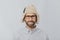 Headshot of cheerful handsome Caucasian guy wears warm winter cap with earflaps, spectacles and white shirt, looks directly at