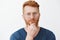 Headshot of creative and smart handsome redhead guy with bristle in glasses and blue t-shirt, rubbing beard on chin and