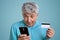 Headshot of mature man with grey hair holds mobile phone and credit card, makes payment online, dressed in blue formal shirt, has