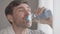 Headshot portrait of perspiring man wiping forehead with hand, drinking refreshing water from bottle and looking at