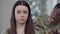 Headshot portrait of sad stressed young woman looking at camera as blurred military man shouting at background. Close-up