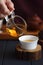 Healthy beverage concept. Pouring freshly brewed tea from glass pitcher into porcelain cup on wood slab on dark background