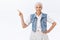 Healthy and joyful, smiling old stylish lady with grey hair, wear trendy denim vest, dress, pointing left, hold hand on