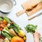 Healthy seasonal vegetables with cooking pot , cutting board and knife on white desk background. Female hand holding fresh bunch