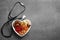 Heart shaped bowl with nuts and dried fruits near stethoscope on grey background, top view.