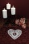 Heart shaped dollie and gemstone, three white candles in metal holders and bouquet of orange and white roses on wooden table.