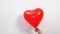 A heart shaped red balloon in hand against a white wall.