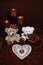 Heart shapped dollie and gemstone, three red candles in metal holoders and red rose, two teddy bears on wooden table.