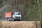 Heavy laden multi-ton truck with white cab and orange body rides along dusty country road in with spring forest background leaving