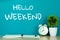 Hello Weekend text on green background and little tree with vintage alarm clock and notebook paper pen on wooden table