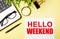 HELLO WEEKEND written on white paper near a computer, magnifier, glasses, pens on a yellow background. Motivational concept. Flat