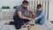 Helpful son is sawing sheet of wood with hand saw while his father is measuring cut with measure-reel and talking to his