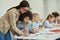 Helpful young female teacher showing how to do task to little schoolboy. Kids sitting at the table, studying in