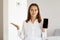 Helpless female wearing white casual style shirt, posing in light room at home, spreading hand aside, looking at camera, showing