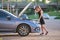 Helpless woman standing near her car with open bonnet inspecting broken motor. Young female driver having trouble with