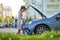 Helpless woman standing near her car with open bonnet inspecting broken motor. Young female driver having trouble with vehicle