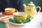 Herbal tea in green cup, honey and yellow teapot on white wooden table in cafe. A cup of hot tea with steam. Natural light