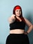 Hey, go on a diet Fat redhead girl in a red headband, black top. She points at you, posing on a blue studio background.