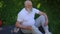 High angle view of old man stroking injured knee sitting on exercise mat on green summer meadow. Portrait of senior