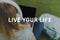 high angle view of young woman sitting on grass with headphones on head and working with laptop, live your life inscription