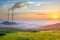 High plant pipes with thick dirty smoke emerging from wide valley with white fog between green mountains