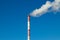 High red and white striped tube lets out white smoke into clear blue sky. Small cloud above pipe on background of electric pole