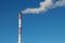 High red and white striped tube lets out white smoke into clear blue sky. Small cloud above pipe on background of electric pole