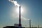 High red and white striped tube lets out white smoke into clear blue sky. Small cloud above pipe on background of electric pole