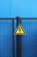 High voltage triangle warning sign mounted on a metal lattice fence on a blue background.